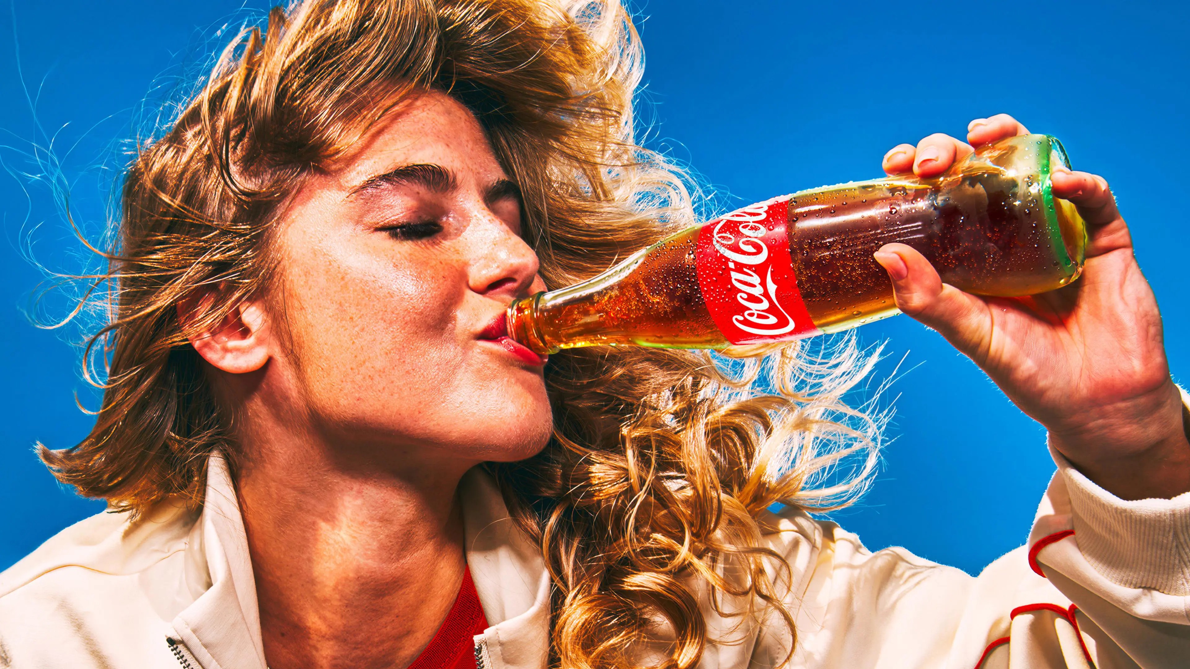 Une femme boit une bouteille de Coca-Cola sur un fond bleu.